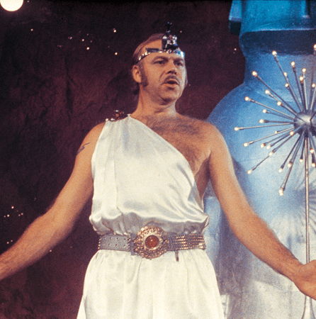 A mid fourties man with a hairy chest in a toga and moustache with arms spread by his side