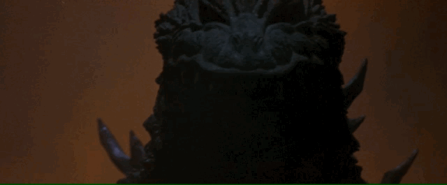Godzilla marches towards the camera, eyes shrouded in darkeness, with burning orange light behind him