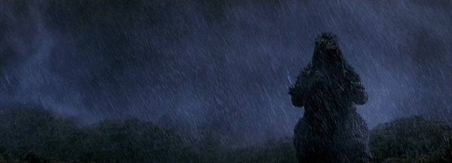 Godzilla marches in the rain, kicks one tank into another, and walks through resulting explosion yelling his signature roar