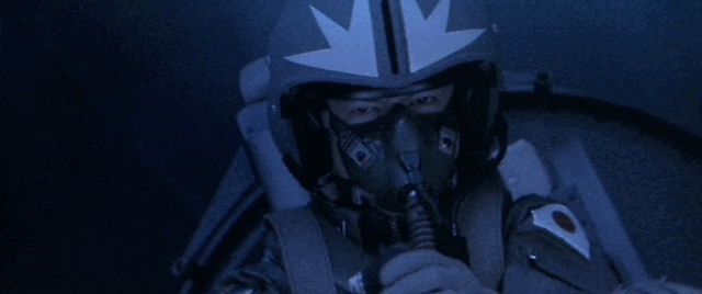 A pilot sits in the cockpit of a jet fighter while Mothra is seen next to it through the clouds